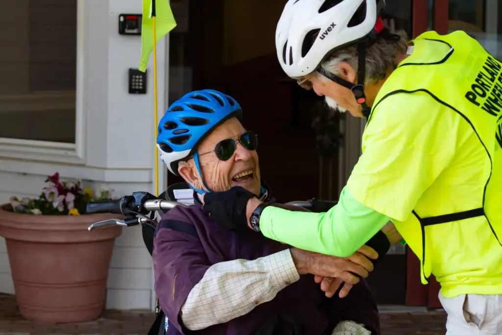A Portland Wheelers volunteer pilot shares a handshake and a laugh with a wheeler. Both pilot and wheeler are white men in their 70s and are wearing bike helmets.
